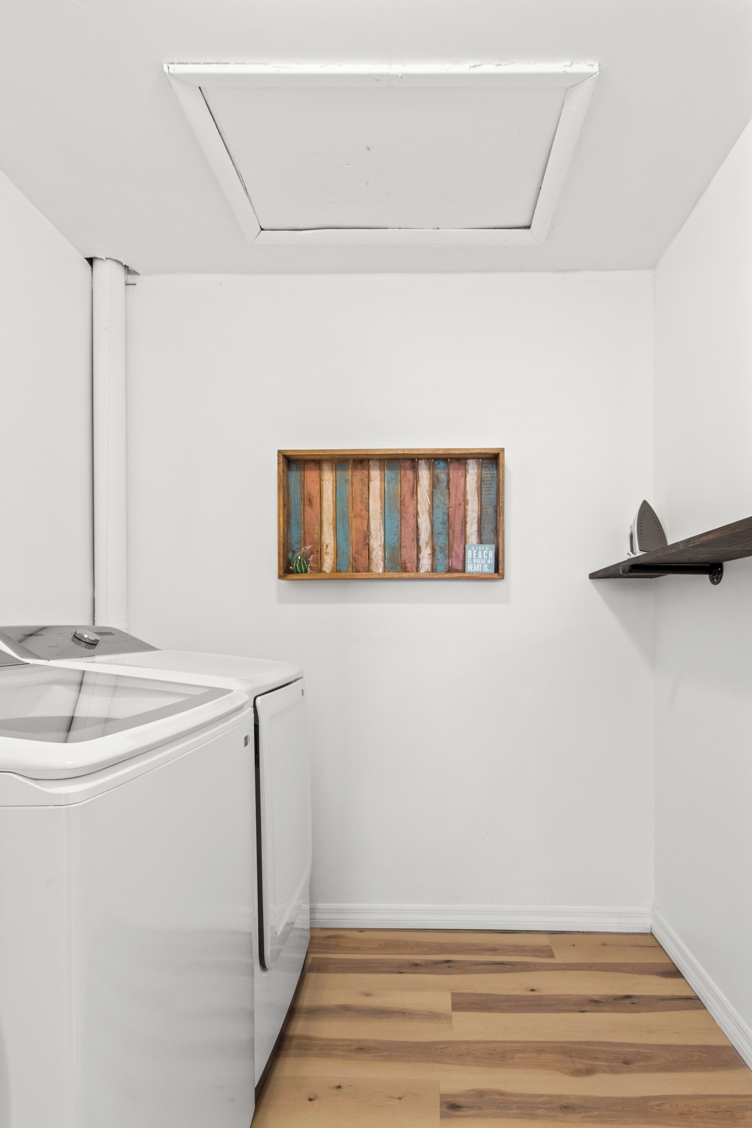 A laundry room with a white washing machine, a wooden-framed artwork on the wall, and a shelf with a surfboard.
