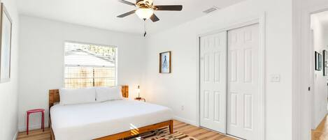 A bedroom with a bed, a ceiling fan, a window with blinds, a small stool, and a patterned rug.