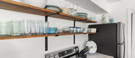 A kitchen with a stainless steel stove, a black refrigerator, and shelves with glassware and dishes.