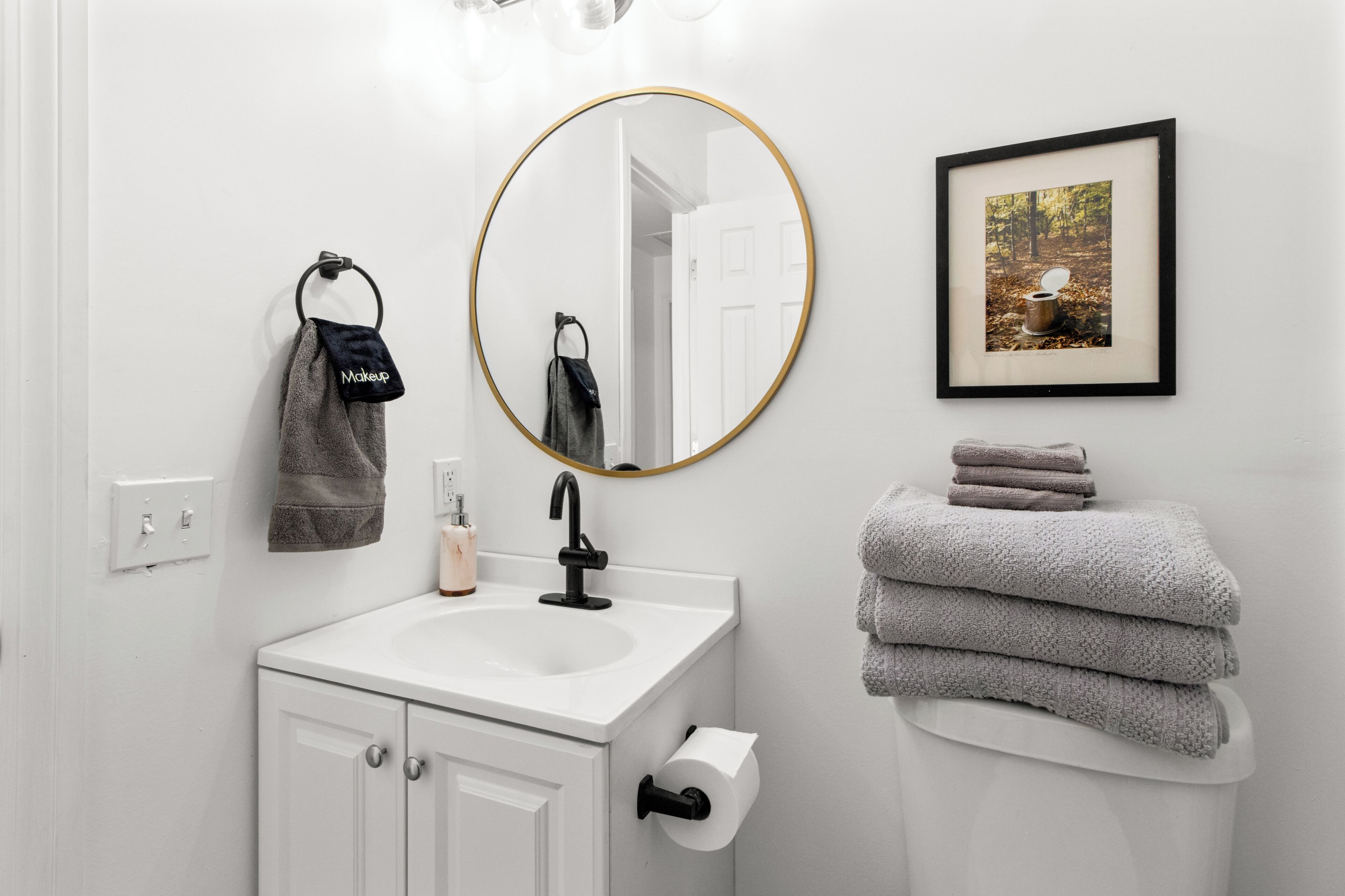 A bathroom with a white sink, a round mirror with a gold frame, a framed picture on the wall, and a stack of towels.