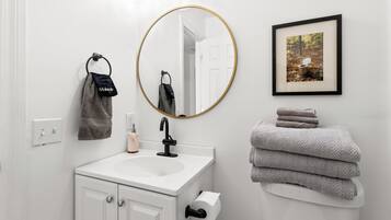 A bathroom with a white sink, a round mirror with a gold frame, a framed picture on the wall, and a stack of towels.