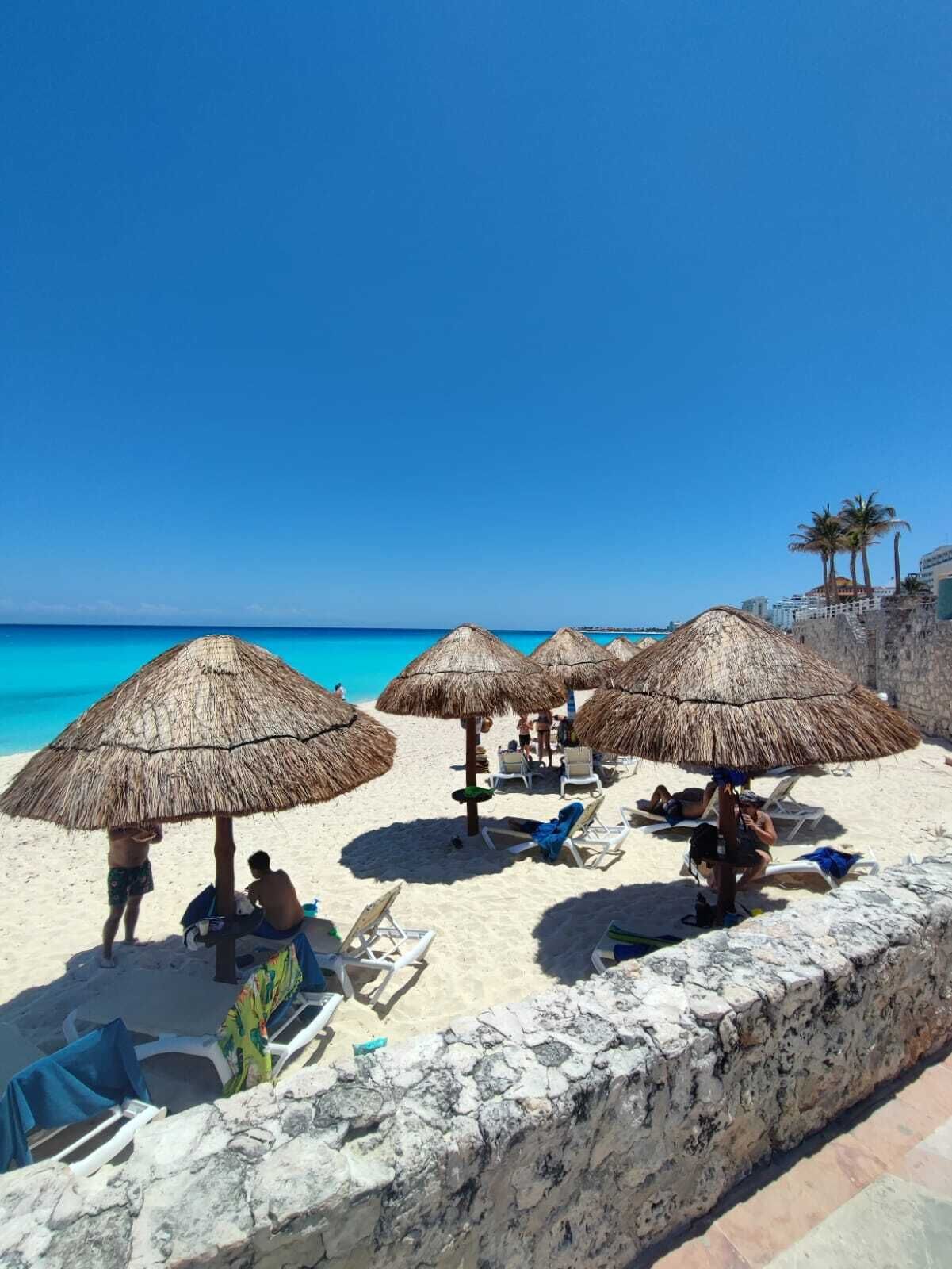 A beach with thatched umbrellas, sun loungers, and a stone wall.