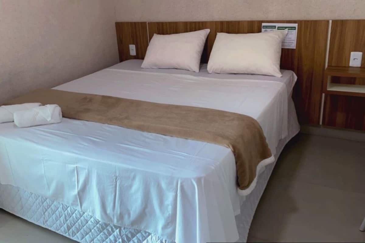 Ein ordentlich bezogenes Bett mit weißer Daunendecke und brauner Tagesdecke, ein Holzkopfschild und ein kleiner Nachttisch mit einer Lampe.