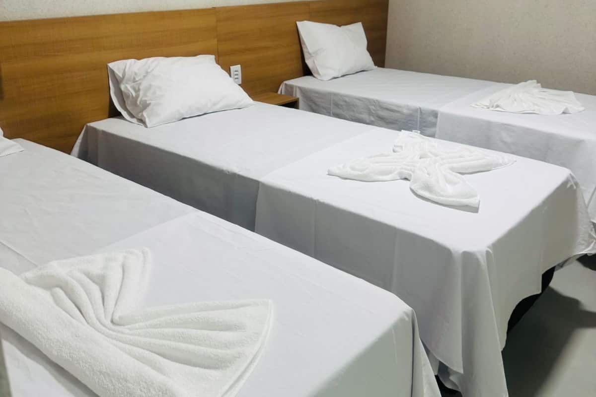Ein Hotelzimmer mit drei Einzelbetten, jedes mit weißer Bettdecke und Kissen. Auf jedem Bett liegen ordentlich gefaltete Handtücher.
