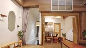 Un salon moderne avec un banc en bois, une petite table et un grand miroir au mur. Une porte donne sur un espace cuisine comprenant une table à manger et des chaises.
