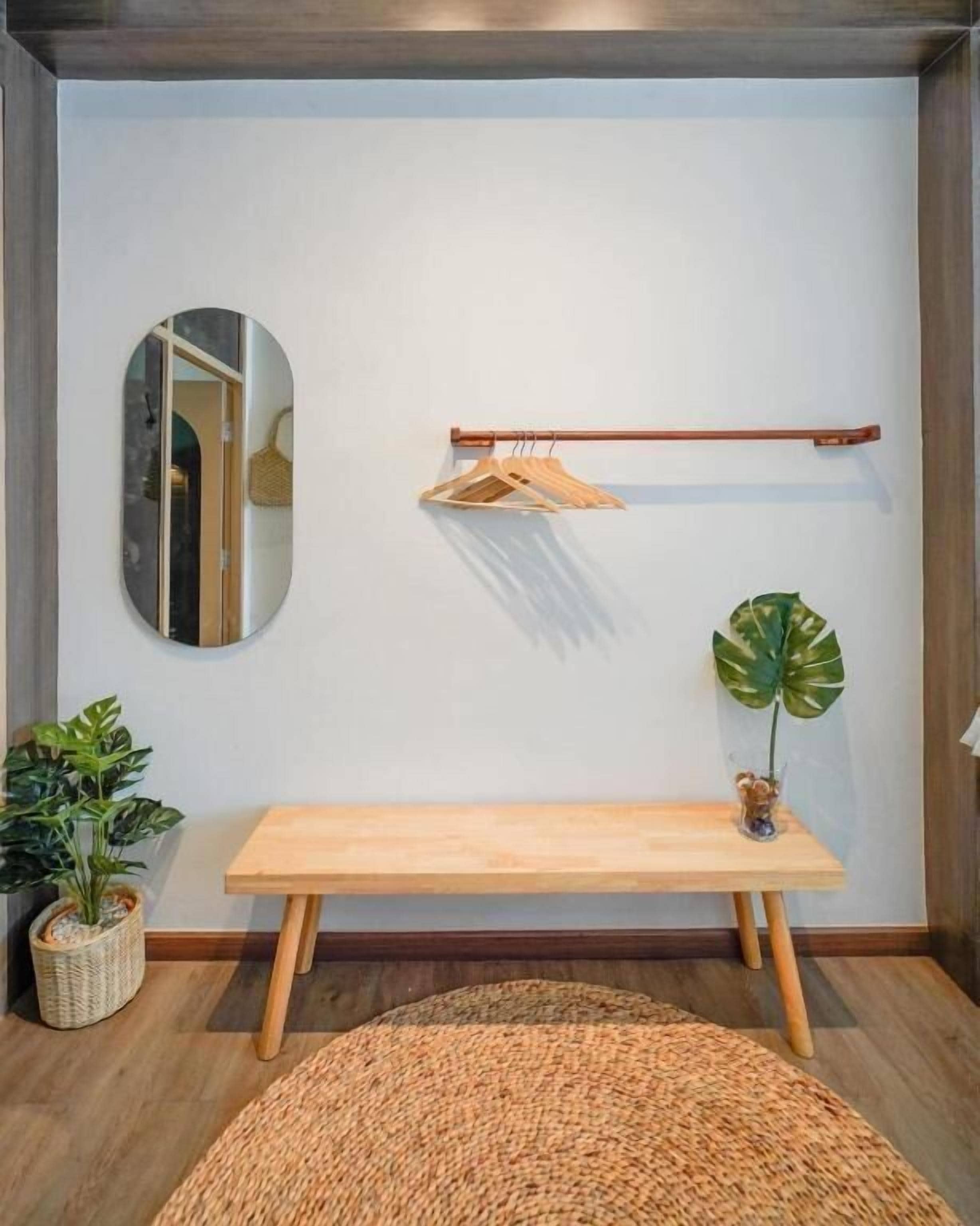A room with a wooden bench, a hanging wooden rack, a round mirror, a potted plant, and a woven rug.