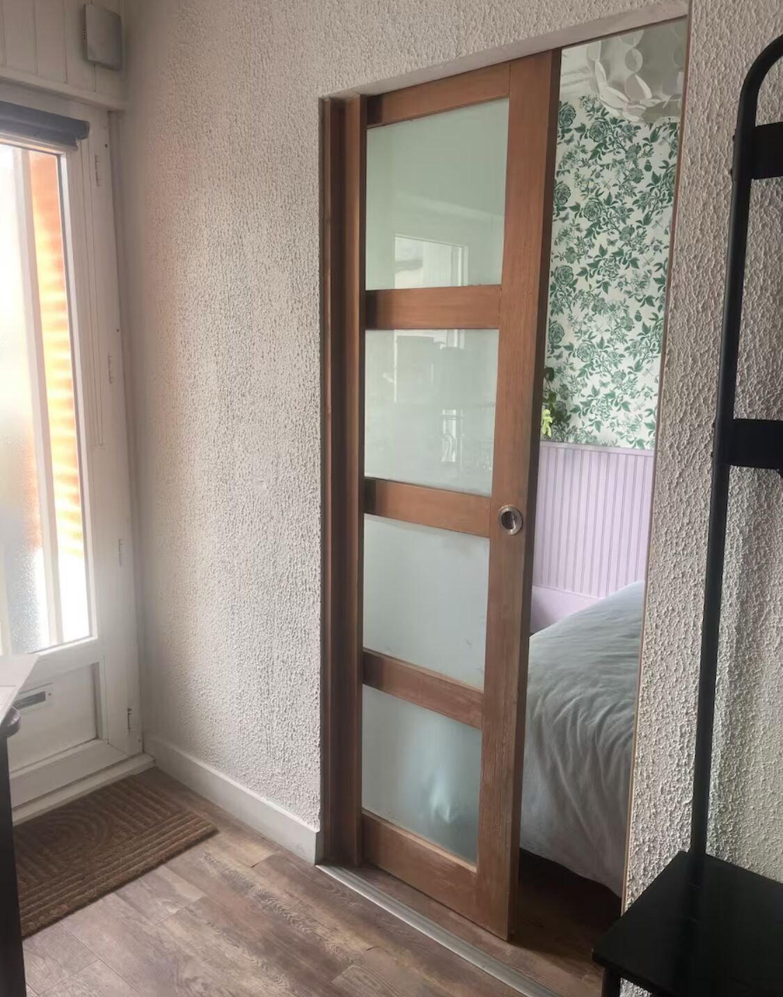 A room with a wooden door with frosted glass panels, a window with blinds, a doormat, and a black metal shelf.