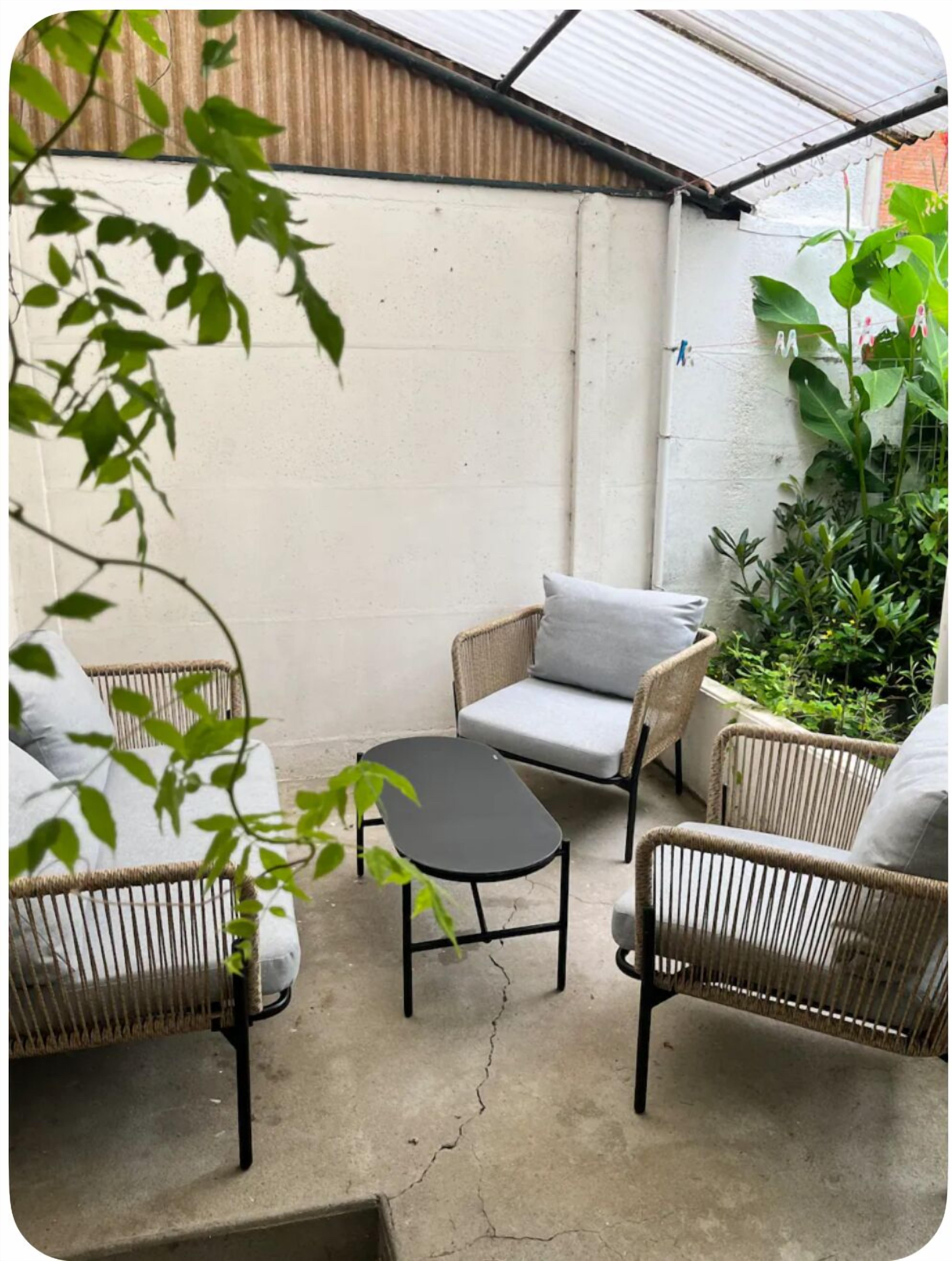 Un pequeño espacio al aire libre con sillas de mimbre y una mesa, rodeado de plantas y un techo de metal corrugado.