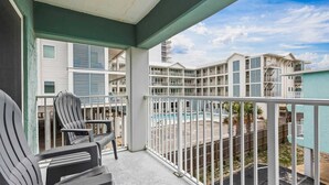 A balcony with two chairs, a pool, and a view of a multi-story building with balconies. - Beachside Comfort | Steps to Sand + Heated Pool (Gulf Shores)