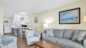 A living room with a grey sofa, patterned armchairs, a wooden coffee table, a dining area with a white table and chairs, a kitchen with white cabinets, and a clock on the wall. - Beachside Comfort | Steps to Sand + Heated Pool (Gulf Shores)