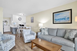 A living room with a grey sofa, patterned armchairs, a wooden coffee table, a dining area with a white table and chairs, a kitchen with white cabinets, and a clock on the wall.
