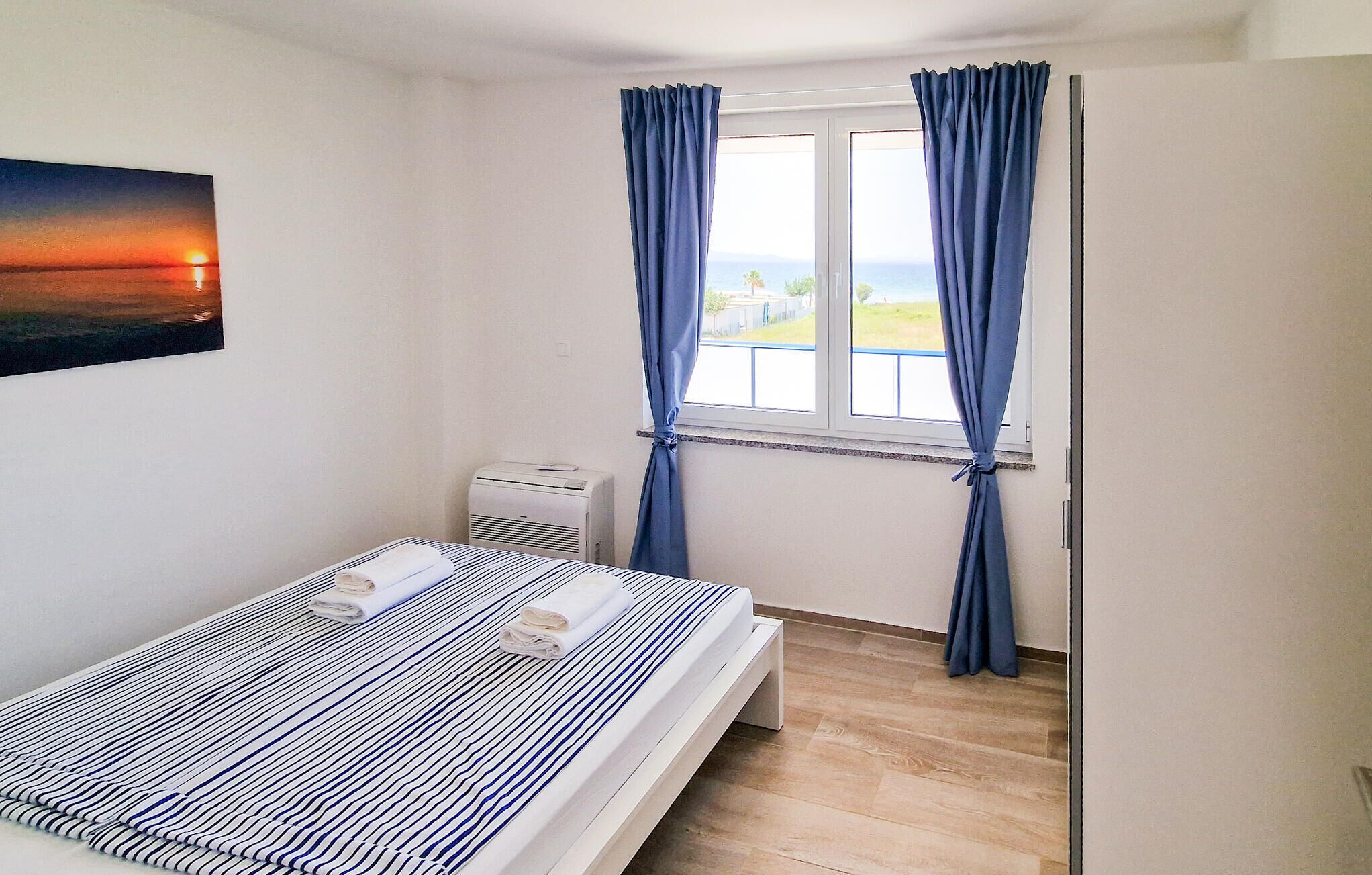 A bedroom with a bed, a radiator, a window with curtains, and a painting of a sunset.