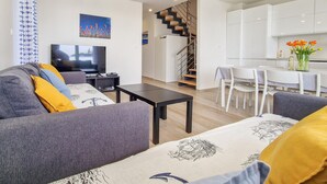 A modern living room with a sofa, coffee table, and TV. There is a staircase leading to a kitchen area with white cabinets and a dining table.