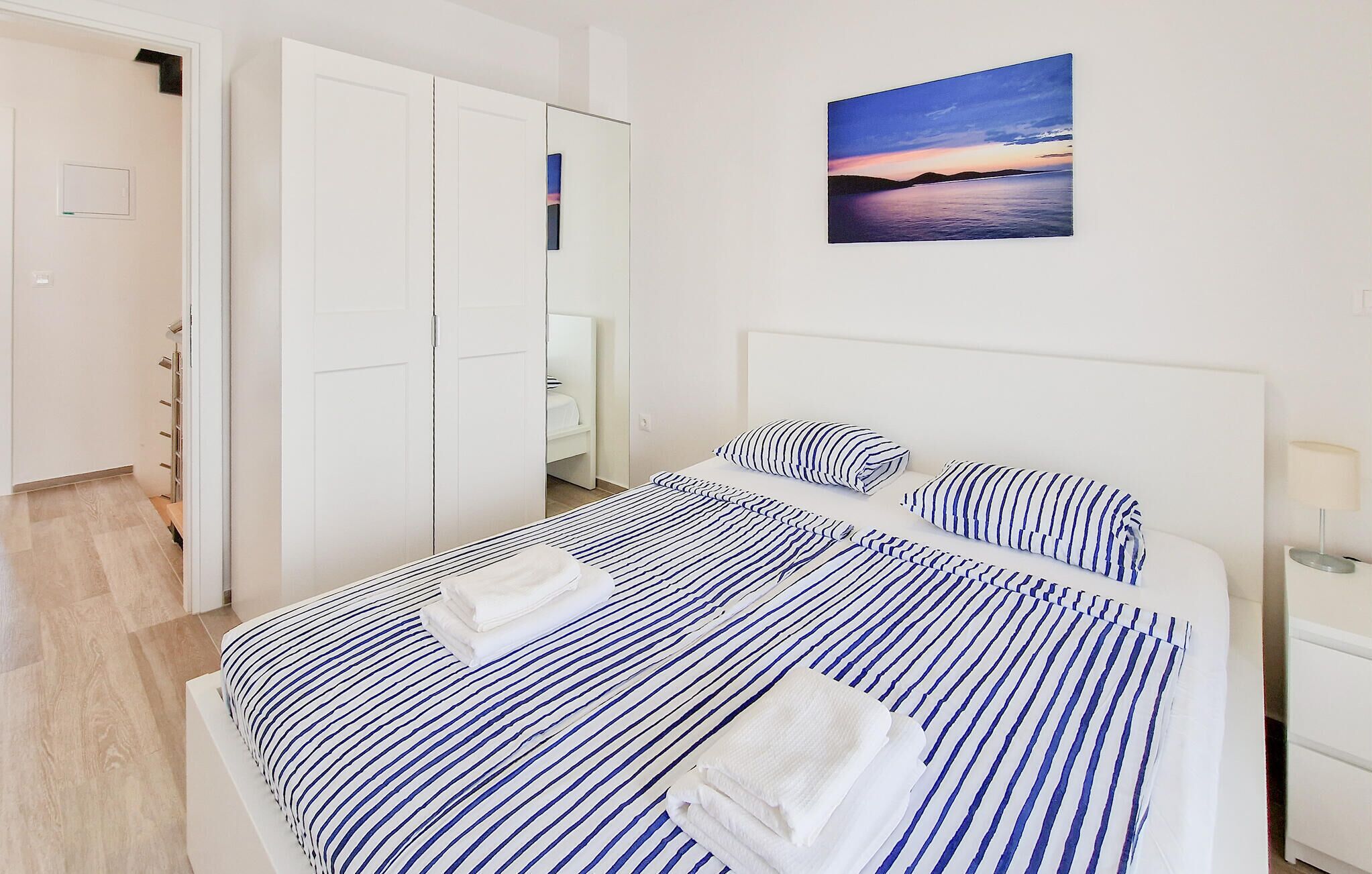A neatly made bed with striped bedding, a bedside table with a lamp, and a painting of a sunset on the wall.