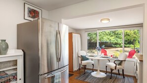 Uma cozinha compacta com geladeira de aço inoxidável, mesa de jantar com quatro cadeiras e vista para o quintal por grandes janelas.