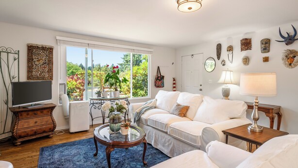 Uma sala de estar com um sofá branco, uma mesa de centro de vidro e um tapete azul. Há uma televisão sobre um suporte de madeira, uma planta em uma mesa lateral e uma janela com vista para vegetação.