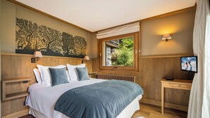 A bedroom with a large bed, wooden headboard, a window with a view of greenery, a bedside table with a lamp, and a television. - 7BR Luxury Chalet with Hammam, Cinema, gym and Mont Blanc Views in Megeve Alps (Megève)