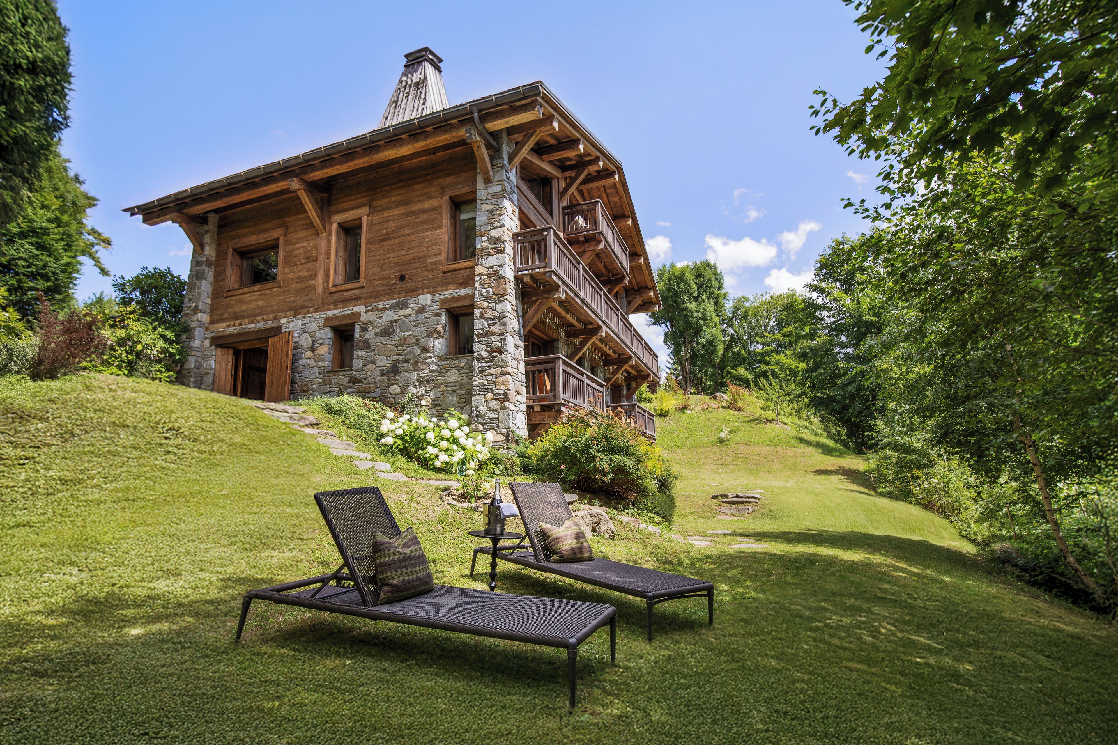 A wooden chalet with a stone foundation, a balcony, and two lounge chairs with cushions.