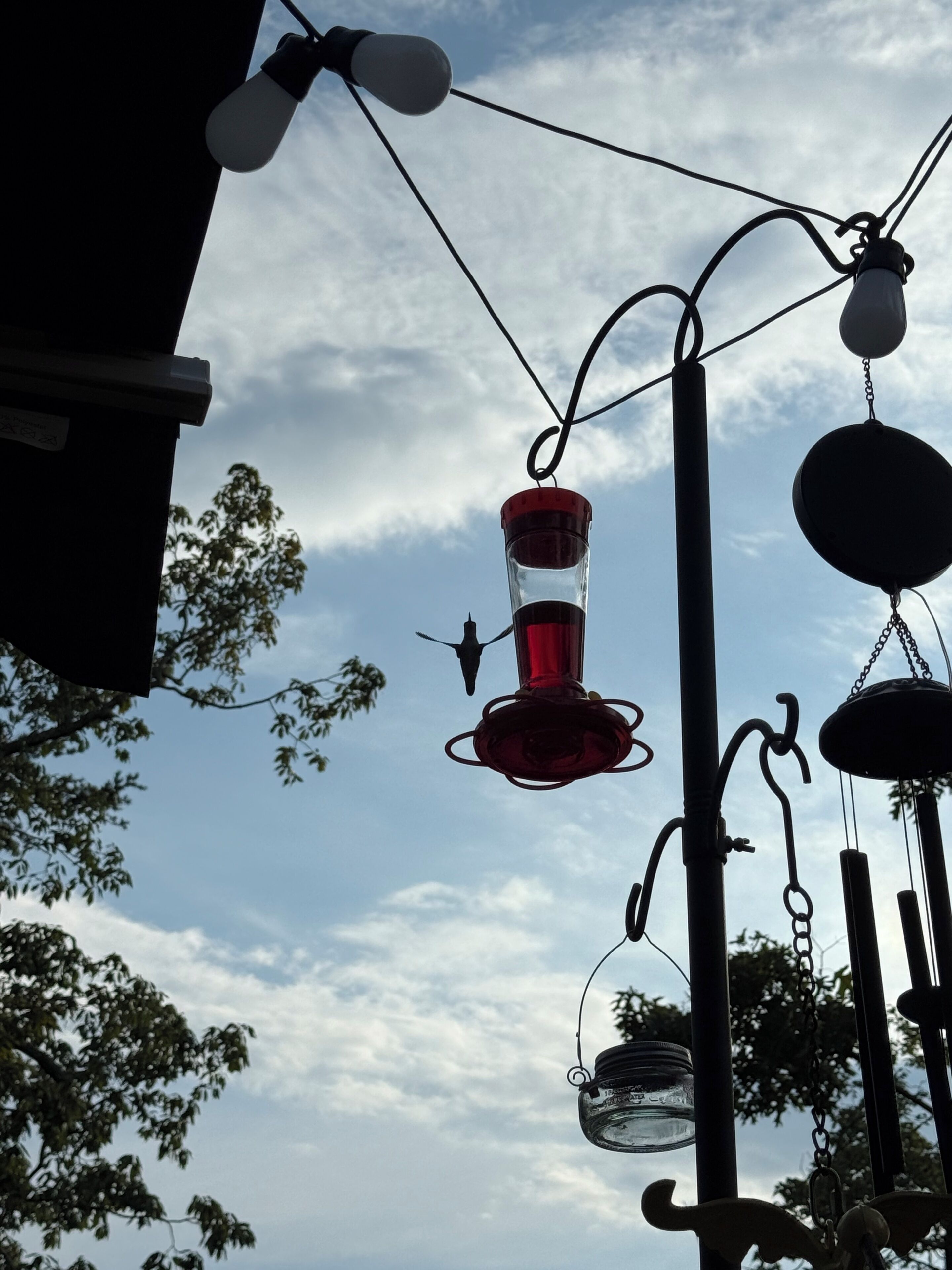 Una lampada a sospensione con una lampadina in vetro rosso e una lampadina in vetro trasparente, appesa con catene metalliche su uno sfondo di cielo nuvoloso.