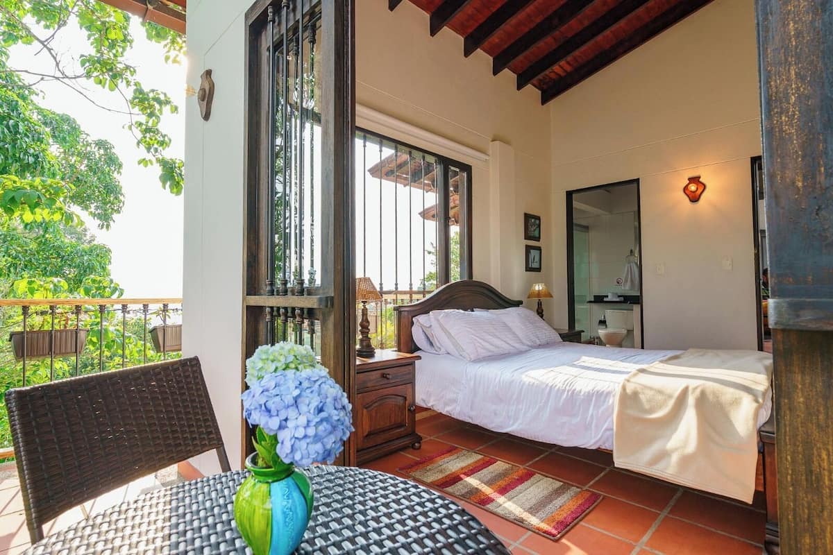 Un dormitorio con cama, un mesita de noche, una mesa con un florero y vistas al exterior.
