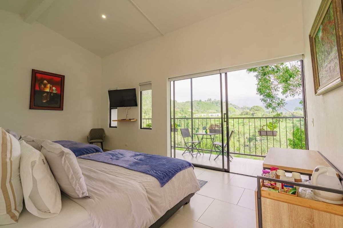 Un dormitorio con cama, televisión, balcón con sillas y vista a la vegetación.