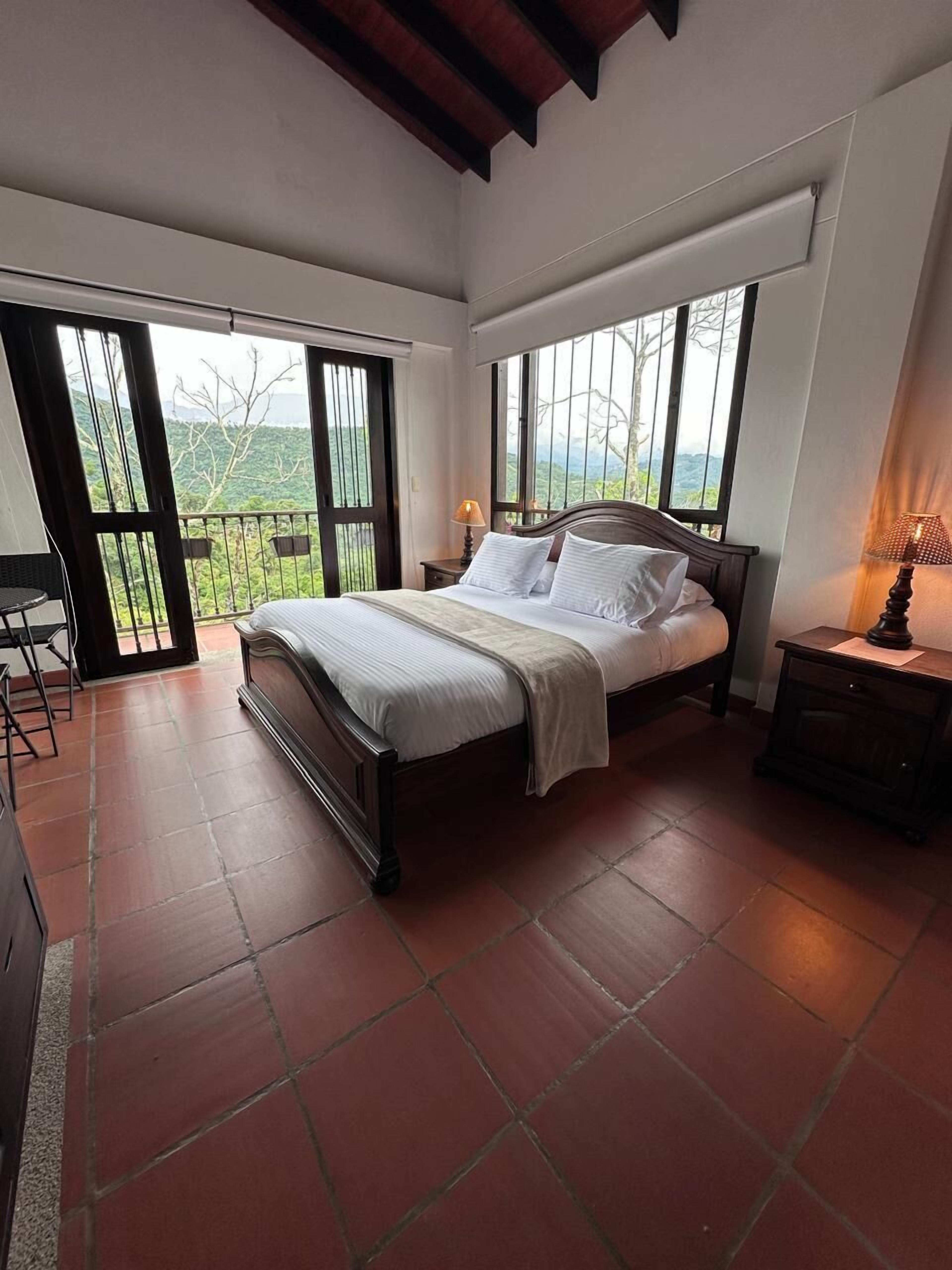A bedroom with a large bed, a nightstand, a chair, and a view of the outdoors through large windows.