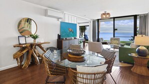 Ein modernes Wohnzimmer mit einem Glastisch, Korbstühlen, einem grünen Sofa und Blick auf den Ozean.