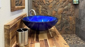 Une salle de bain avec un lavabo en verre bleu, un meuble-lavabo en bois, un mur en pierre et un espace douche.