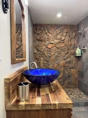 A bathroom with a blue glass sink, wooden vanity, stone wall, and a shower area.