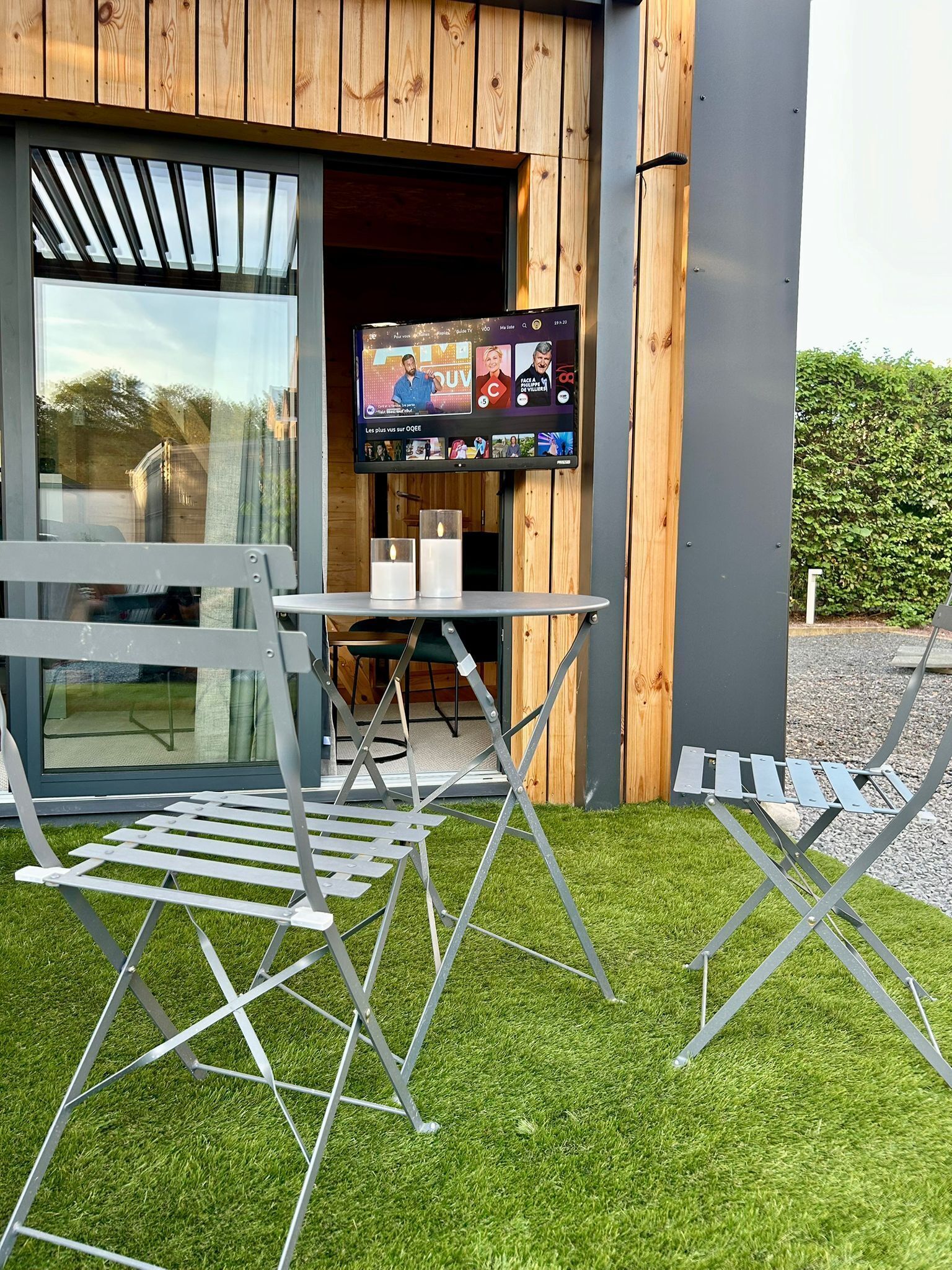 An outdoor seating area with a table and chairs, a flat-screen TV mounted on a wall, and a wooden building facade.