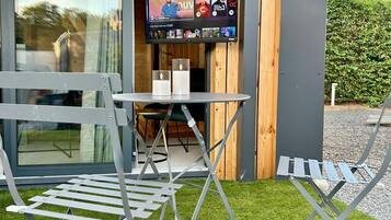 An outdoor seating area with a table and chairs, a flat-screen TV mounted on a wall, and a wooden building facade.
