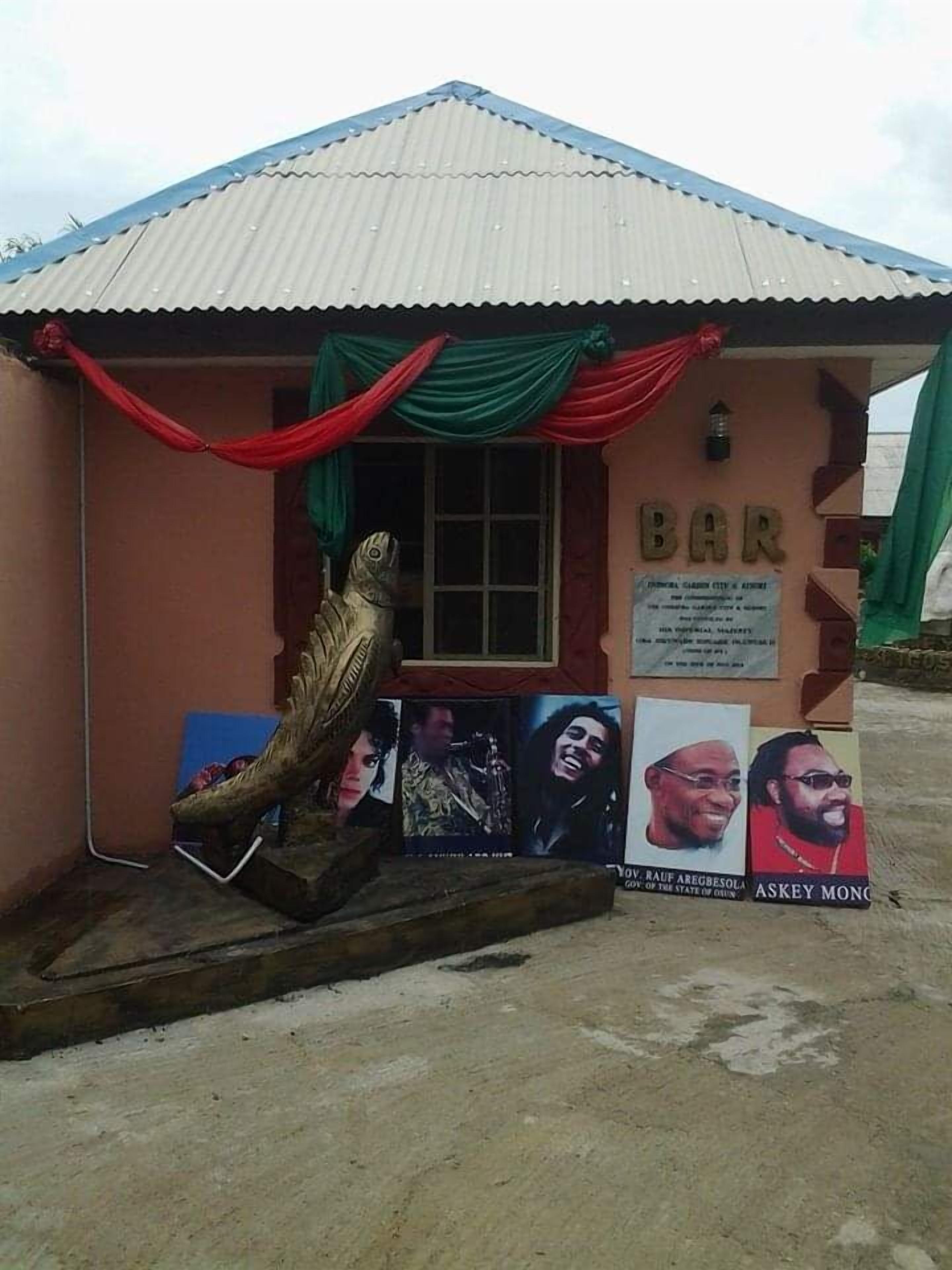 A small building with a corrugated metal roof, a window, and a sign that reads "BAR". There are posters with blurred faces and a wooden sculpture of a fish.