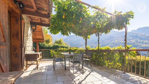 Ein Holzhaus mit einer Steinwand, einem Balkon mit Tisch und Stühlen und Blick auf eine Berglandschaft.