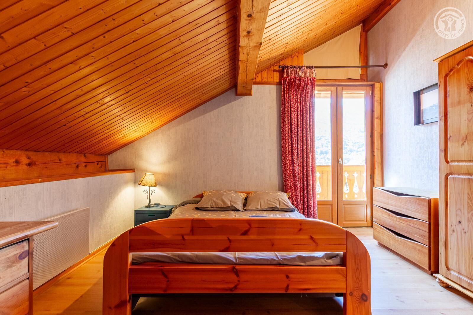 Une chambre à coucher avec un plafond en pente, un lit en bois, une table de chevet, une lampe et une vue sur l’extérieur, visible à travers une porte.