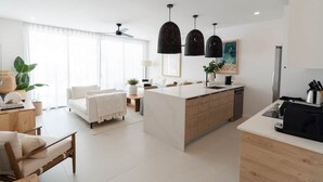 Una cocina moderna con una isla central, iluminación colgante y un área de comedor con sofá y mesa blancos.