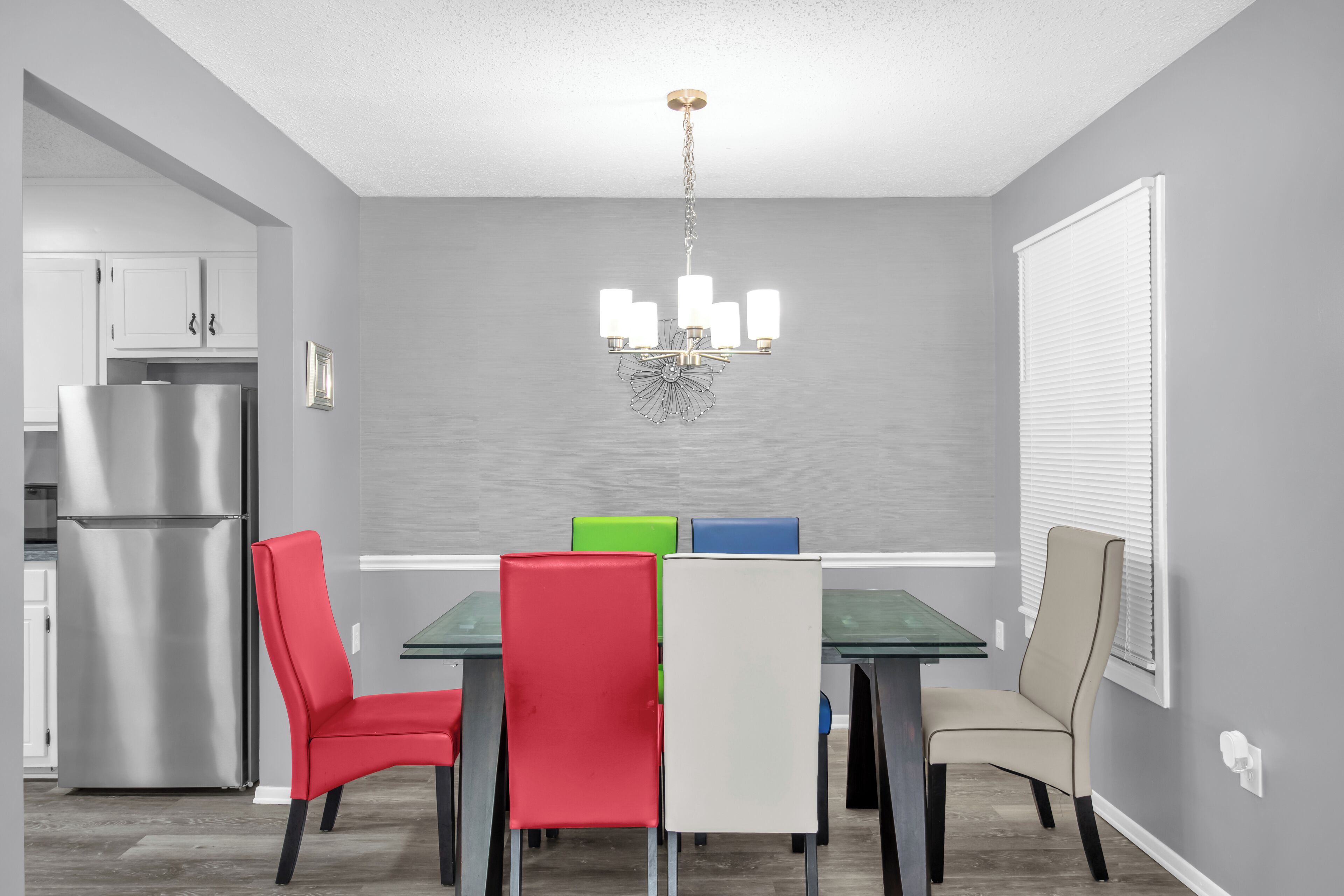 A dining area with a glass table, four chairs, a chandelier, and a refrigerator in the background.