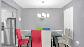 A dining area with a glass table, four chairs, a chandelier, and a refrigerator in the background.