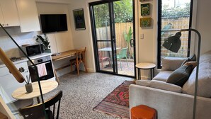 A modern kitchen with a microwave, stove, and sink, a dining area with a table and chairs, a sofa, and a large sliding glass door leading to a patio with outdoor furniture. - Stylish Studio Apartment with Private Courtyard (Newtown)