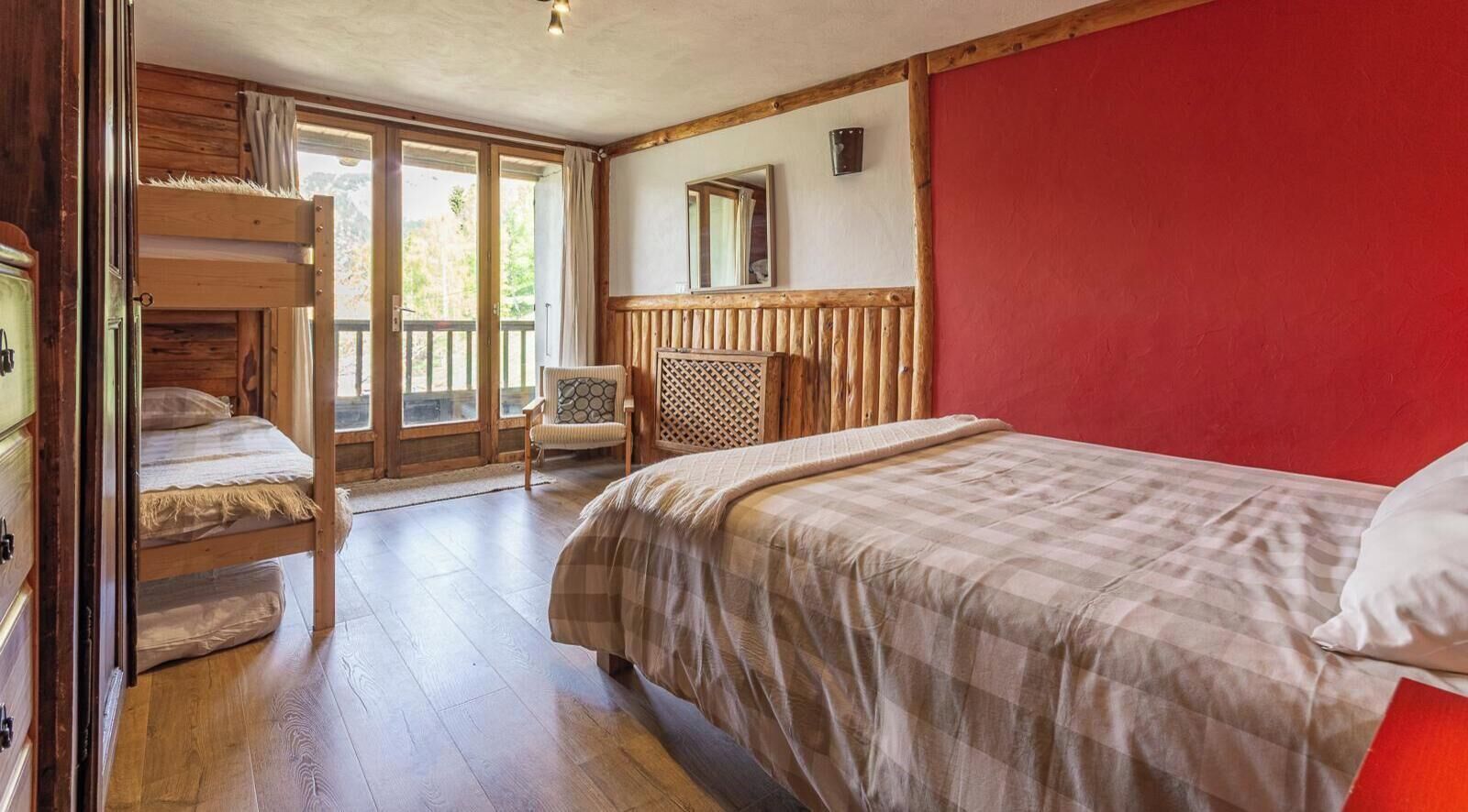 Ein Schlafzimmer mit einem Etagenbett, einem Einzelbett, einem Stuhl, einem Holz-Kopfteil und einer roten Akzentwand.