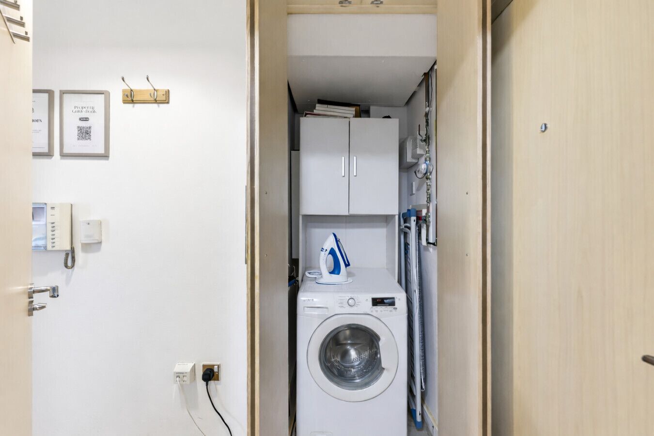 A compact laundry room with a washing machine, a dryer, and a small ironing board. There are framed pictures on the wall and a telephone on the left.