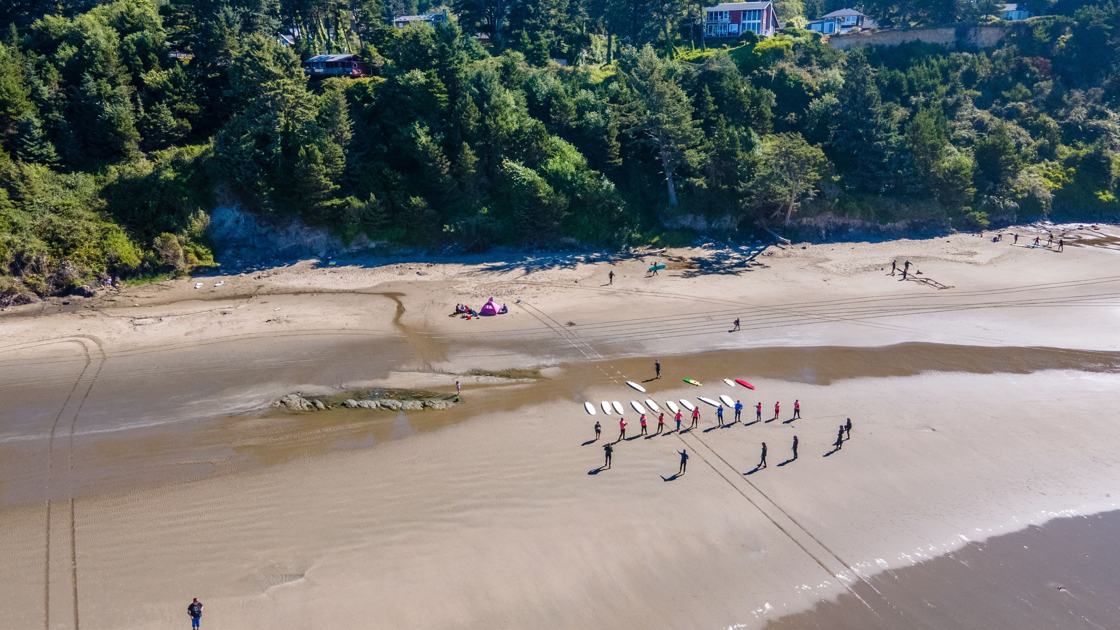 Aerial view of a sandy beach bordered by forested hills, with surfers gathered on the shore.