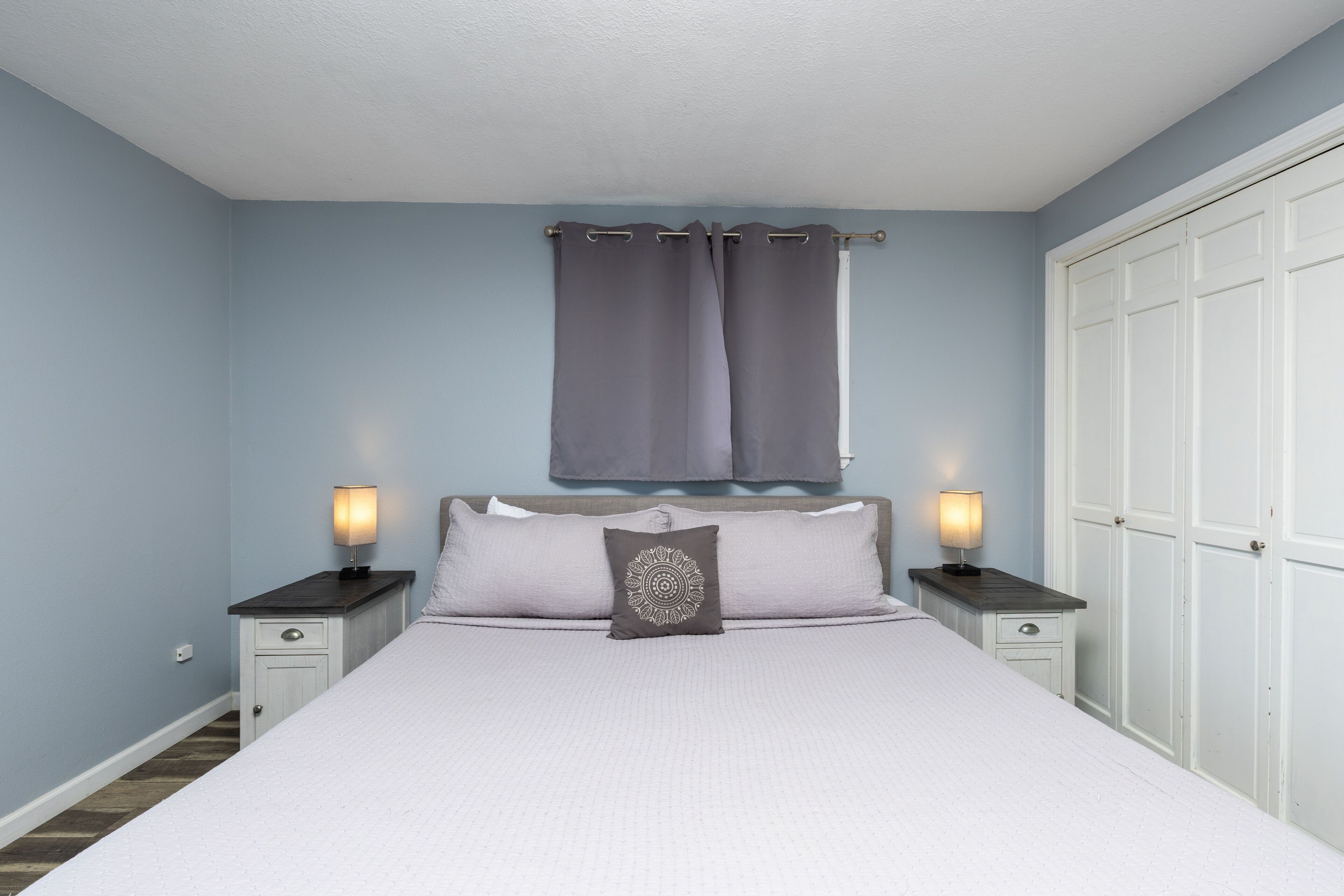A bedroom with a bed, two bedside tables with lamps, a nightstand, a window with curtains, and a wardrobe.