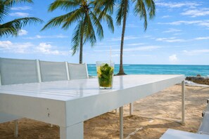 A white table with a glass of drink on it, set on a sandy beach with palm trees and the ocean in the background.