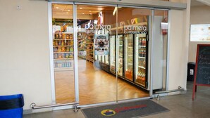 Entrada de un supermercado con puertas de vidrio, un pasillo visible en el interior y un letrero de pizarra en el exterior.