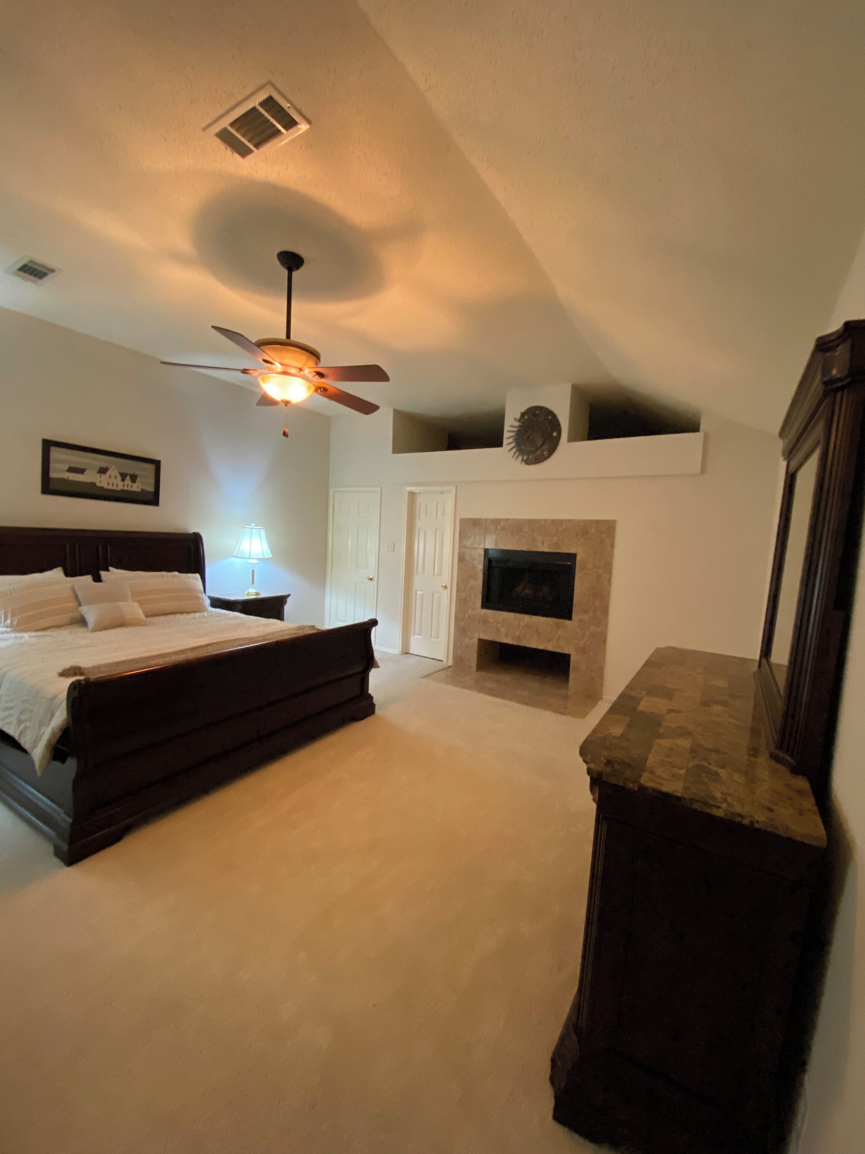 Un dormitorio con cama, ventilador de techo, chimenea y mesita de noche con lámpara.