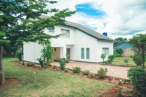 Une maison à deux étages avec une allée pavée, un petit jardin agrémenté de diverses plantes et un grand arbre au premier plan.