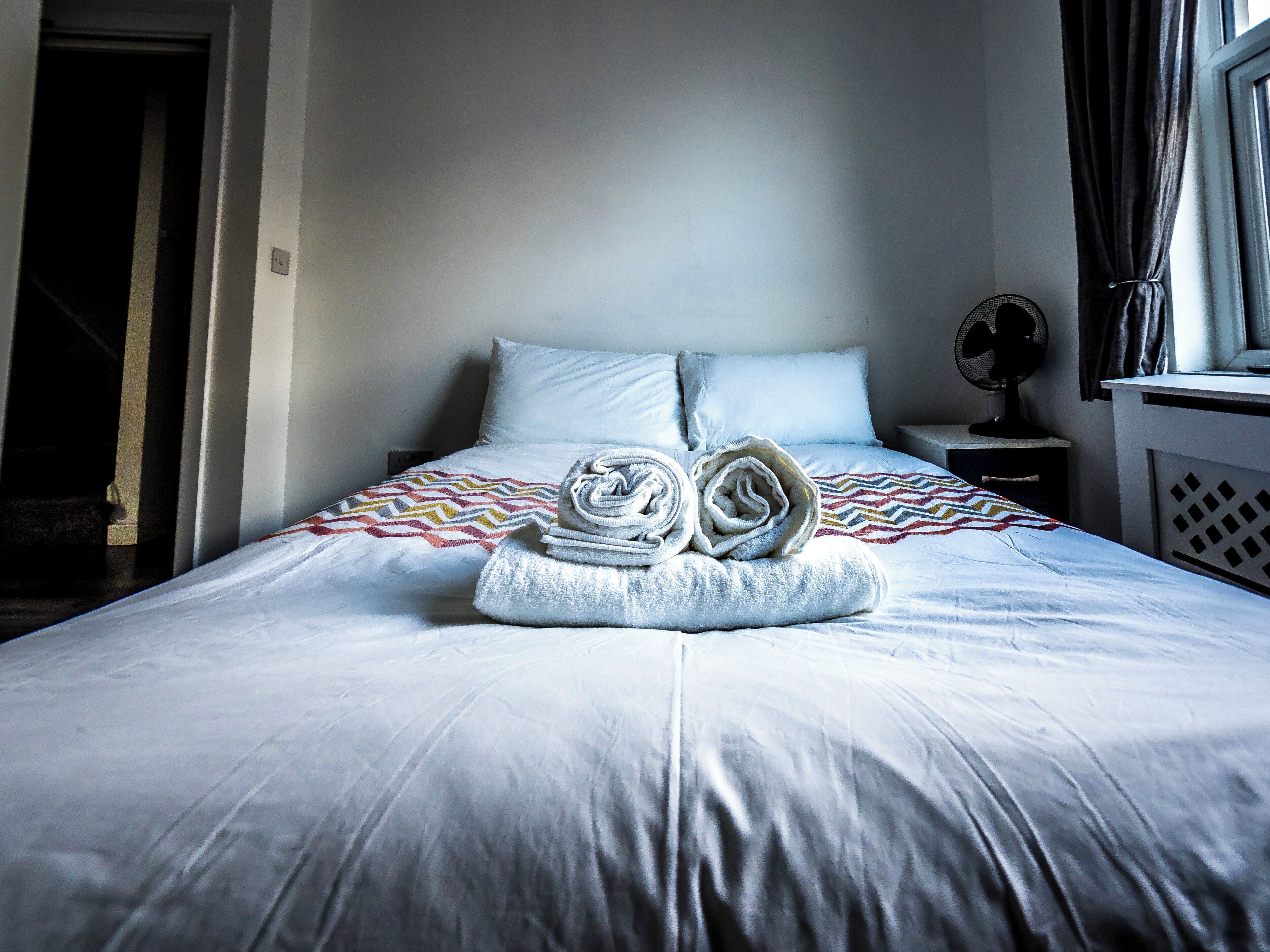 Cama bem arrumada com um cobertor estampado, uma toalha dobrada e um ventilador em cima da mesa de cabeceira.