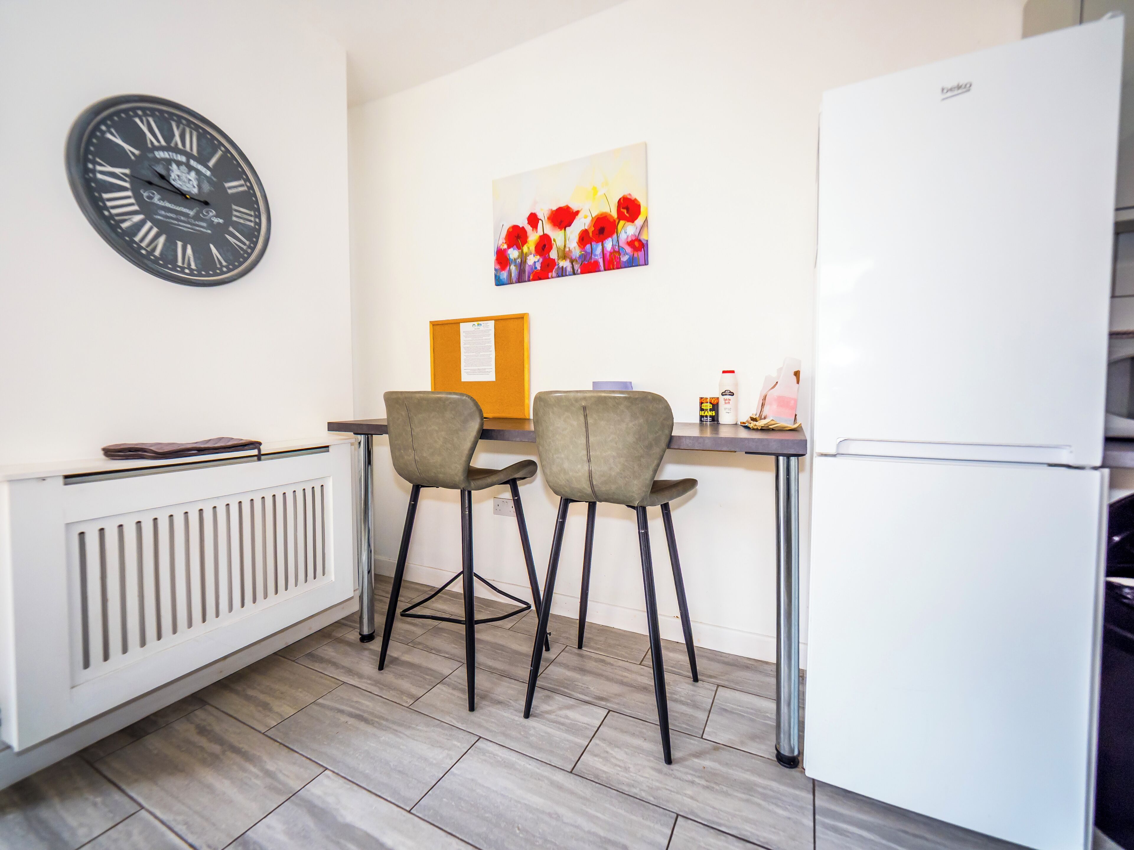 A small kitchenette with a white refrigerator, a clock, two chairs, a table, and a bulletin board.