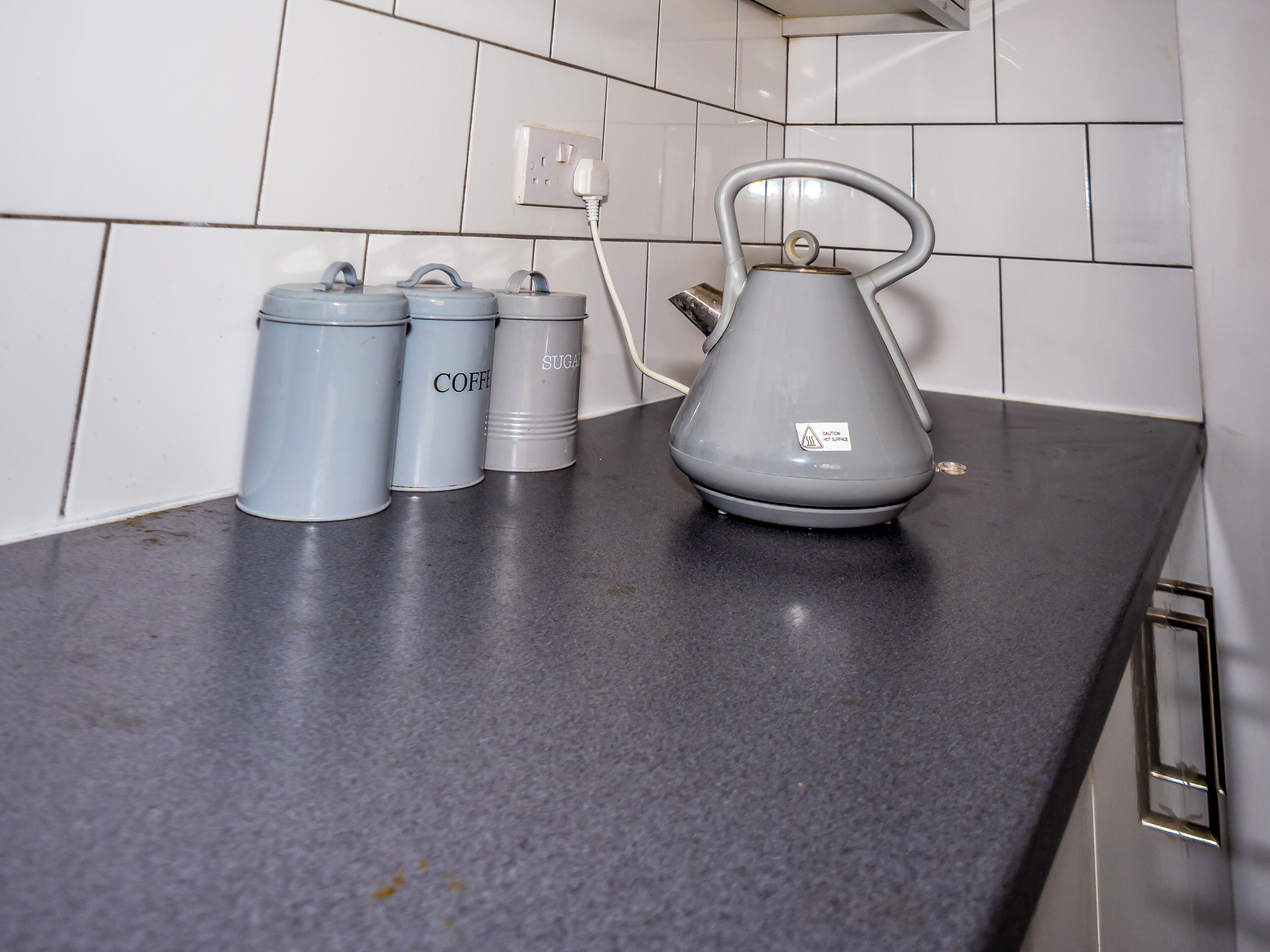 A kitchen counter with a kettle, two containers labeled "coffee" and "sugar," and a power outlet.
