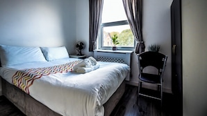 Un letto rifatto con un copriletto fantasia, un comodino con una ventola, una finestra con tende e una sedia.